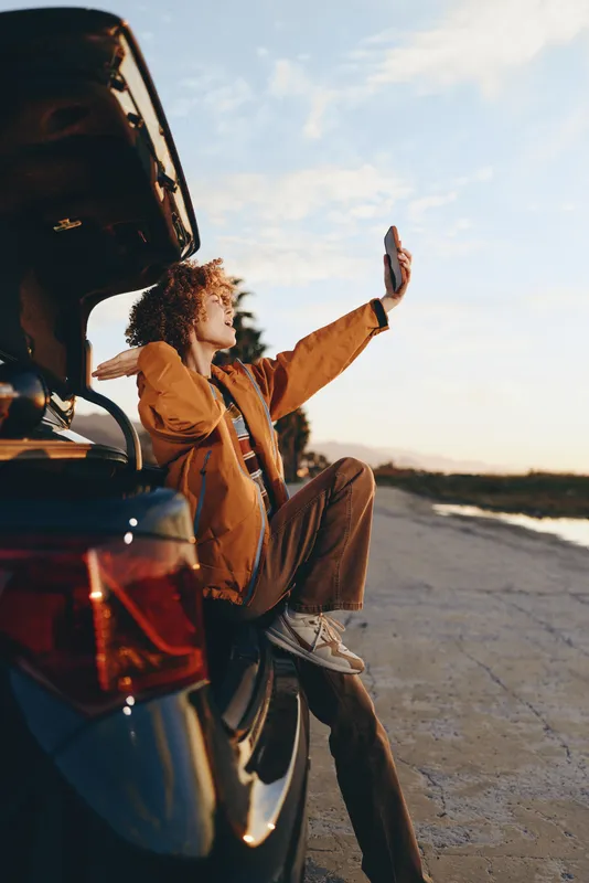 Woman lifestyle near an open car trunk on the road at sunset. She poses with one arm raised, warm sunlight and outdoors travel and adventure journey atmosphere.