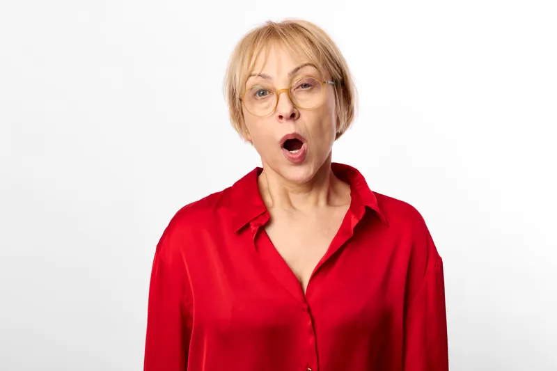 Elderly woman portrait with surprise expression, mouth open, wearing red shirt, looking at camera, clean white studio background, soft focus, direct gaze