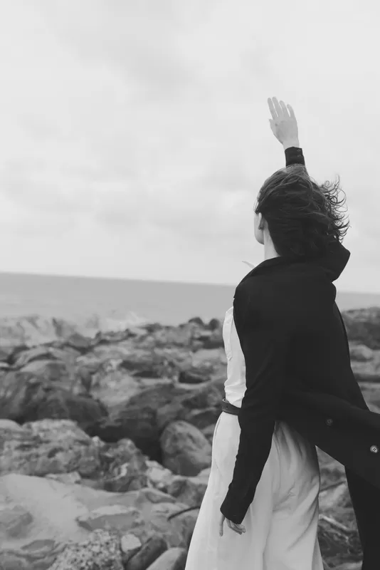 Black and white scene of a lone figure on a rocky shore raising an arm toward the sea, wind blowing, dramatic mood and coastline in monochrome, cinematic mood bold, dramatic.
