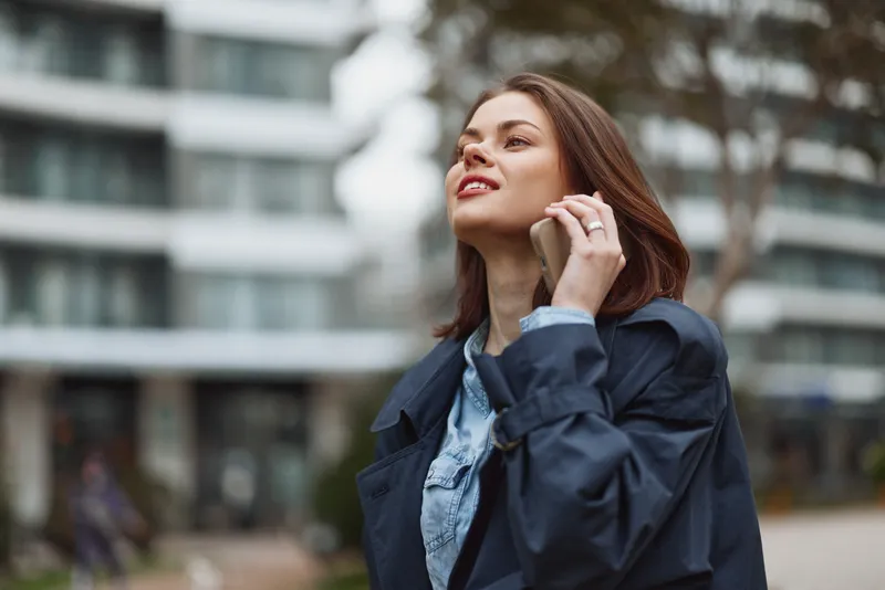 Smiling woman in a navy jacket talks on her phone outdoors in an urban park. Bright daylight highlights casual fashion, confidence, and everyday communication.
