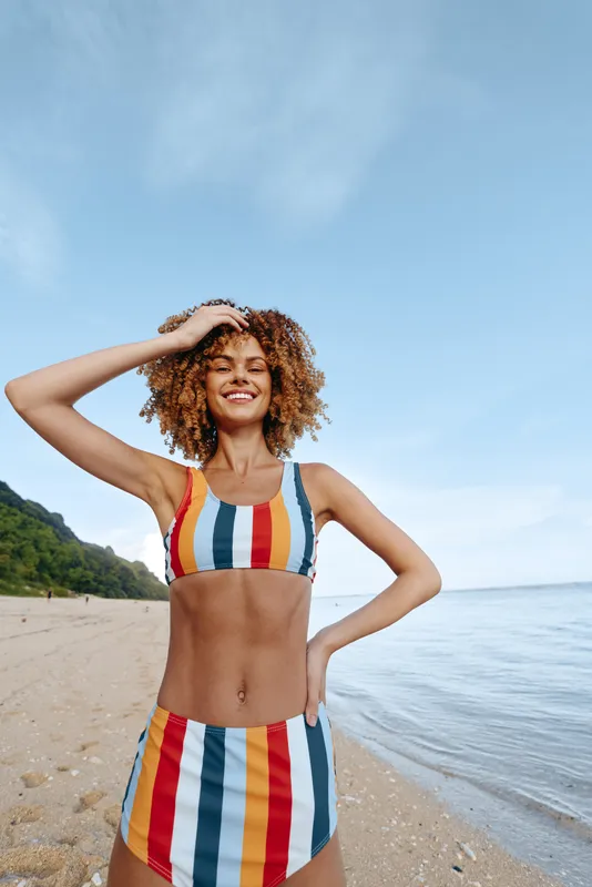 Young woman with curly hair in a colorful striped swimsuit smiles happily while standing on a sandy beach near calm sea water under a clear sky.