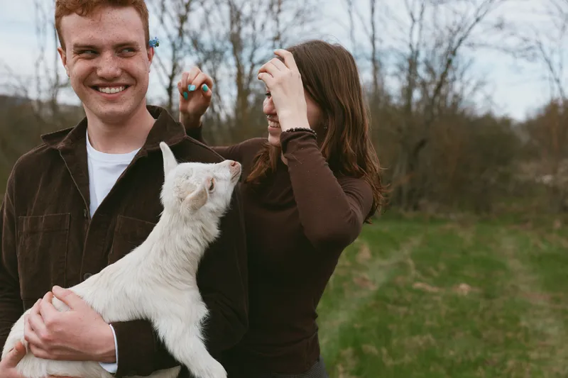 Cheerful couple outdoors with a friendly goat kid, warm smiles, gentle interaction, natural lighting, grassy field, candid moment, relaxed mood, everyday life setting, wholesome countryside atmosphere