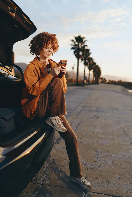 Woman lifestyle car phone sunset road travel casual palm outdoors, young woman leans on parked car, checking smartphone, smiling, in jacket and jeans on open road at sunset