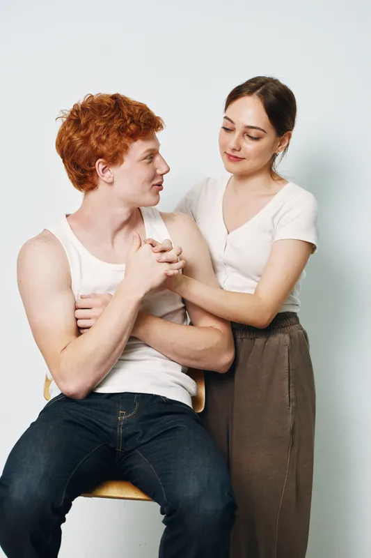 Couple, young man and woman, sitting and standing, holding hands and looking at each other with love, emotions and connection in casual clothing indoors.