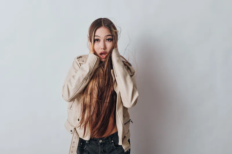 Young woman with long hair and surprised expression wearing light jacket and dark jeans posing against plain background for fashion, emotion and casual portrait.