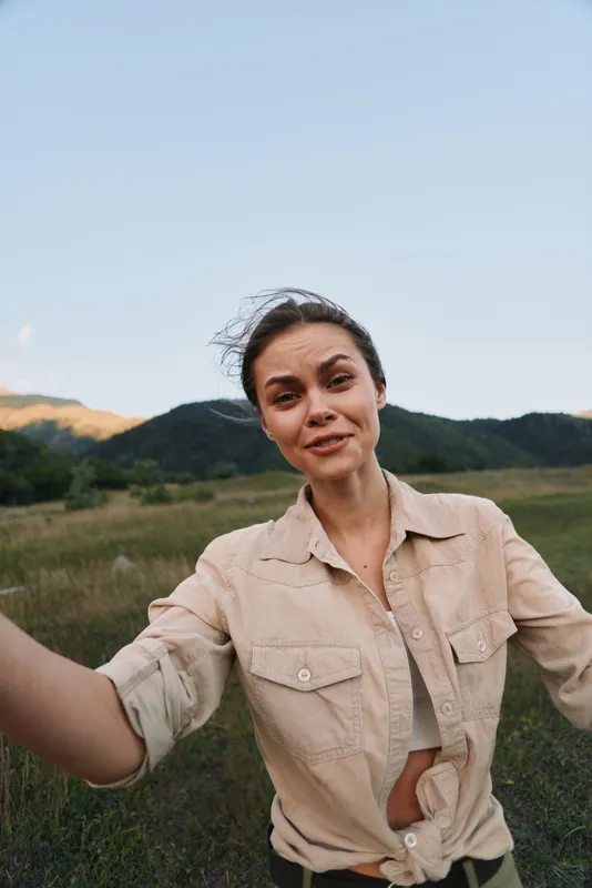 Young woman outdoors in a natural field takes a casual self portrait, wearing a light beige shirt, smiling softly, warm sunlight, relaxed lifestyle mood, close framing.