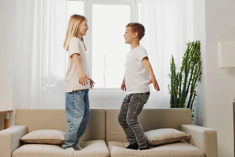 Two happy children jump on a beige sofa in a bright living room with large windows and a tall green plant. Casual clothes and playful mood reflect childhood energy and fun at home.