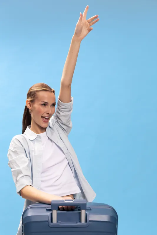 Happy young woman with long hair stands behind blue suitcase, raising hand and smiling on blue background. Travel, vacation, and joyful concept for trip preparation and excitement.