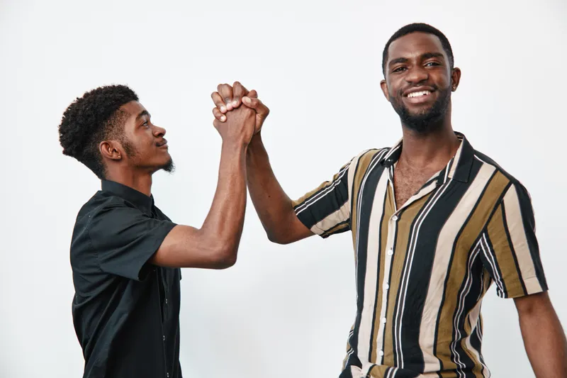 Two young black men with dark skin shaking hands and smiling in casual clothes on white background. Friendship, connection, and positive interaction between African ethnicity males.