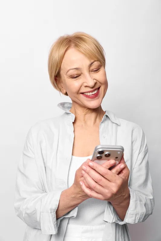Older woman smiling while holding smartphone, close portrait on white background, relaxed posture, technology and communication, everyday lifestyle, soft studio light
