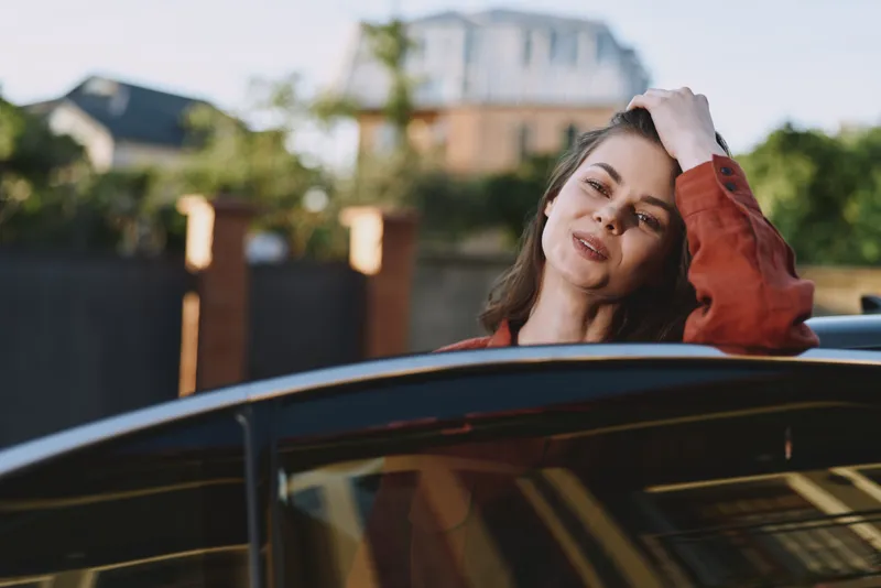 Young woman leans on a sleek vehicle, smiling softly on a sunny day with a relaxed pose, urban background, breeze in hair, casual style