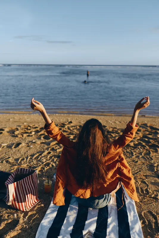 Beach ocean woman arms raised at sunset, horizon and sky above calm waves, warm sunlight on sand, curly hair and denim jacket, joyful vacation moment, standing carefree, outdoors