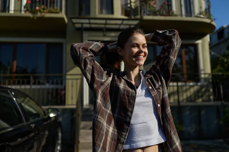 Casual outdoor portrait of a smiling woman wearing a plaid shirt, enjoying daytime sunlight, relaxed pose in front of a residential building, lifestyle mood