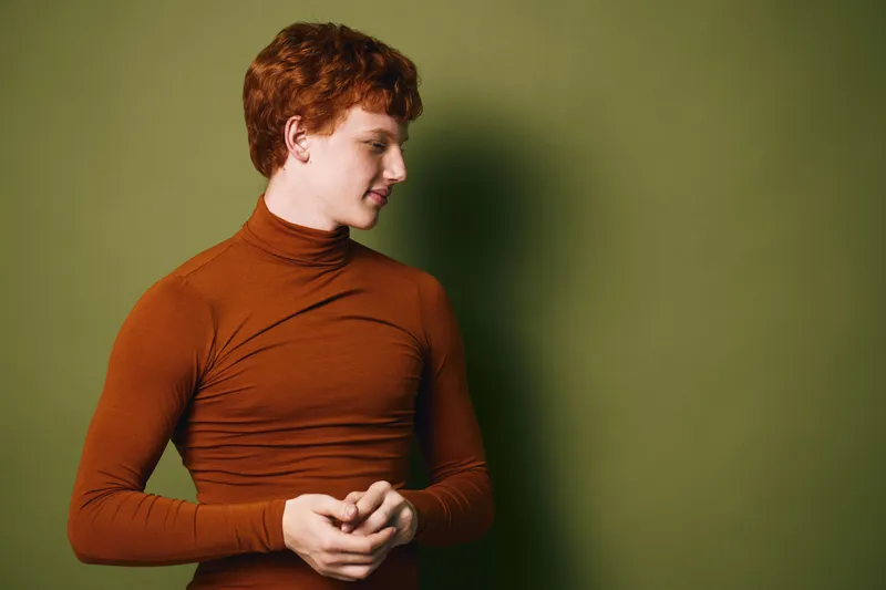 Young man with red hair and orange turtleneck sweater standing against green background with hands clasped and head turned to side in thoughtful pose for portrait.