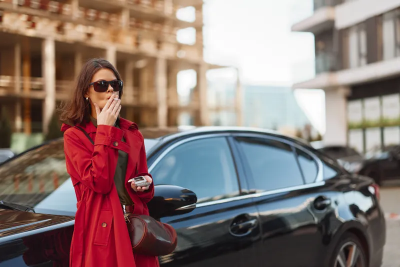 Young woman in a red coat and sunglasses stands beside a black car in an urban setting while covering her mouth with her hand, expressing surprise or thoughtfulness.