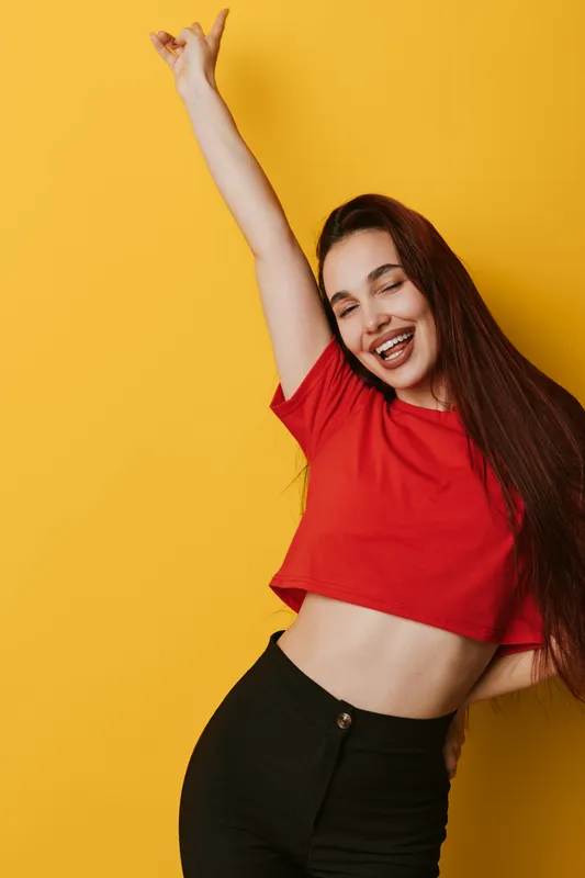 Young woman with long hair wearing red crop top and black pants smiling with eyes closed and raising arm against yellow background for happiness and celebration concept.