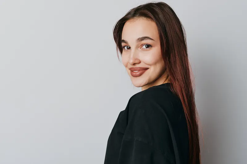 Young woman with long brown hair and natural makeup smiling and looking over shoulder against gray background. Casual style, confident expression and relaxed pose in studio portrait.