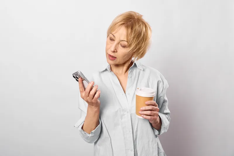 Concerned woman holds smartphone and coffee cup, looking at screen, indoor studio portrait, white background, casual shirt, copy space, relaxed posture near wall.