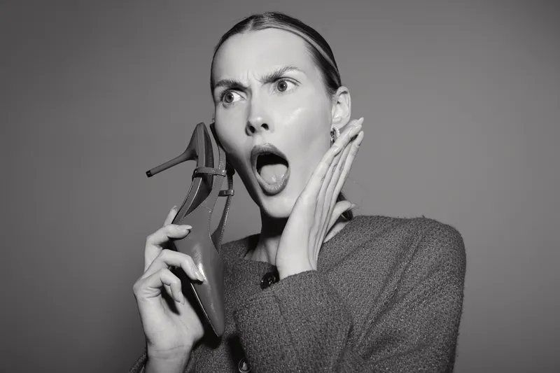 Shocked white woman with light skin holds high heel shoe near face, mouth open and eyes wide. Black and white studio portrait captures emotion of surprise and disbelief concept.