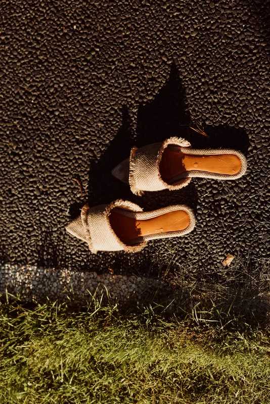A pair of beige slip-on sandals with fluffy straps rest on a textured asphalt surface casting long shadows in warm natural light.