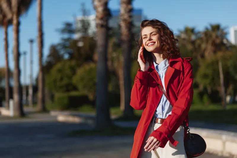 Smiling woman in a red coat talking on a cellphone outdoors. Sunny park setting with palm trees, chic fashion vibe, candid lifestyle moment with warmth