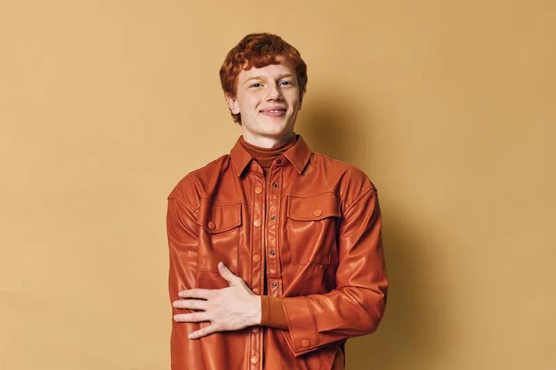 Young man with red hair wearing orange leather jacket standing with crossed arms against beige wall smiling confidently in casual outfit for fashion portrait lifestyle concept.
