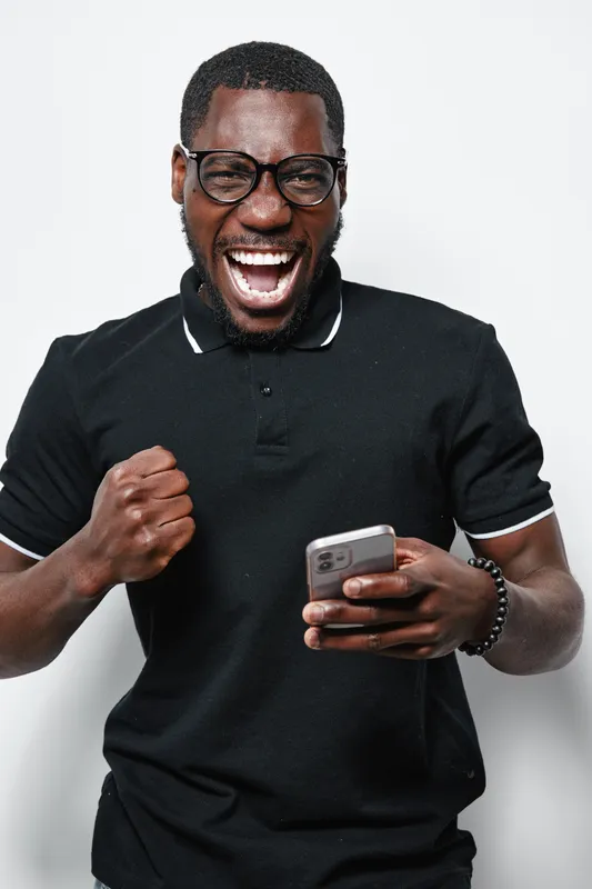 African American man with dark skin wearing glasses and black polo shirt holding smartphone and celebrating success with fist pump and joyful expression on white background.