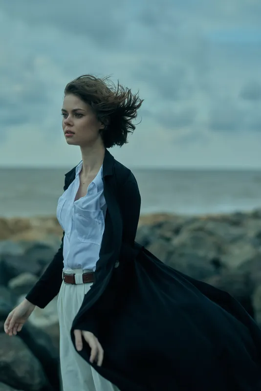 Windy seashore scene with a figure walking along the rocky coast, flowing coat, light shirt, and expansive ocean background creating a moody, atmospheric sense of motion and elegance