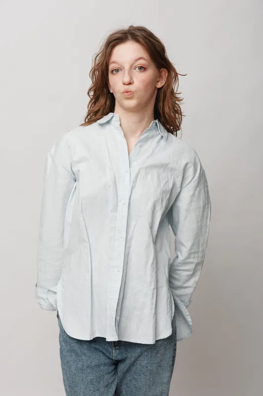 Young woman in white shirt and blue jeans makes funny face with pursed lips, standing against plain light background. Casual style and playful mood concept.