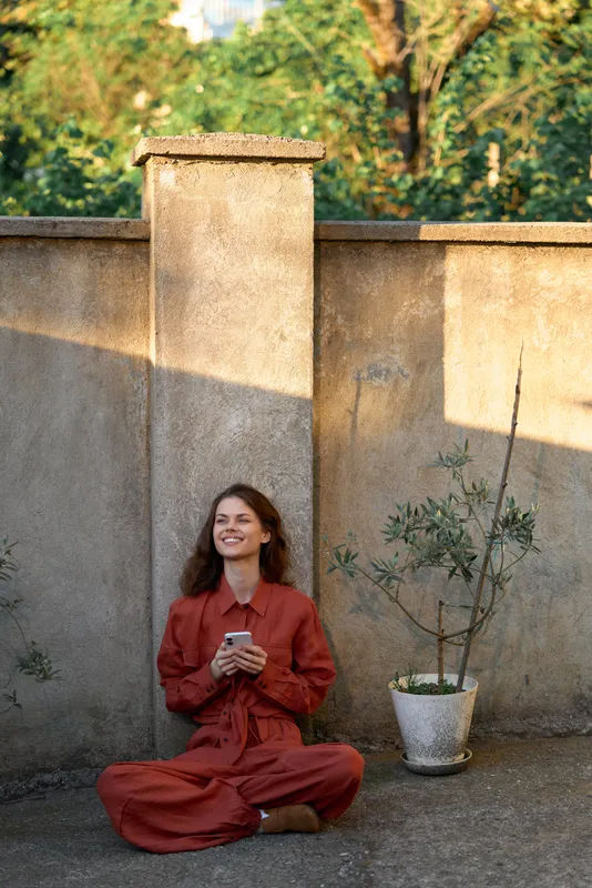 Woman in a red outfit sits outdoors, smiling while using a smartphone. Warm sunlight, rustic wall, and a potted plant create a relaxed lifestyle moment of calm