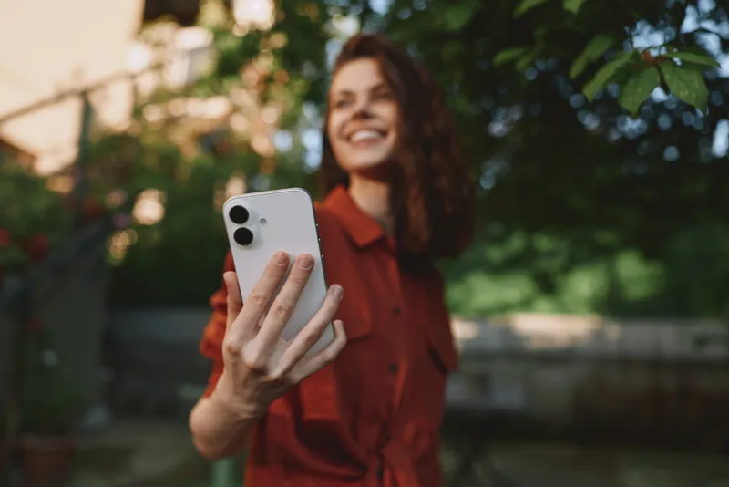 Smiling woman outdoors taking a selfie with a smartphone, wearing a red top, in a sunny park, lifestyle and self expression, casual mood and natural light. Soft, warm ambiance