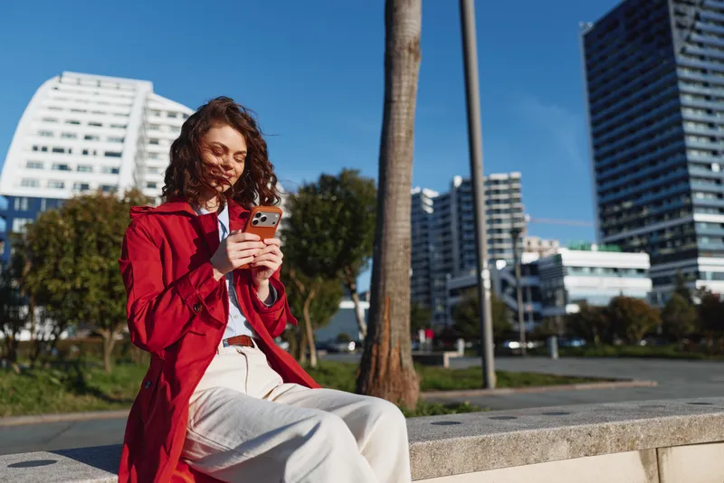 Young woman in a red jacket sits outdoors on a sunny day while smiling at her smartphone in a modern urban environment.