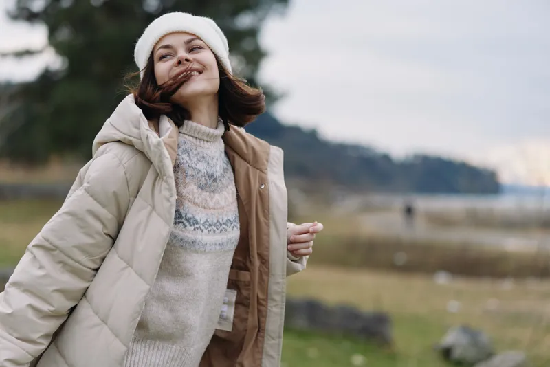 Woman in a cozy winter coat smiles outdoors, wearing a knit sweater and beanie, with a quilted jacket in a windy landscape by a lake with trees and open sky
