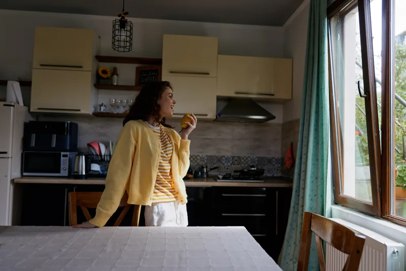 A young woman in a yellow cardigan stands in a bright kitchen, taking a bite of an apple and enjoying a casual morning, her relaxed pose suggesting a simple everyday moment.