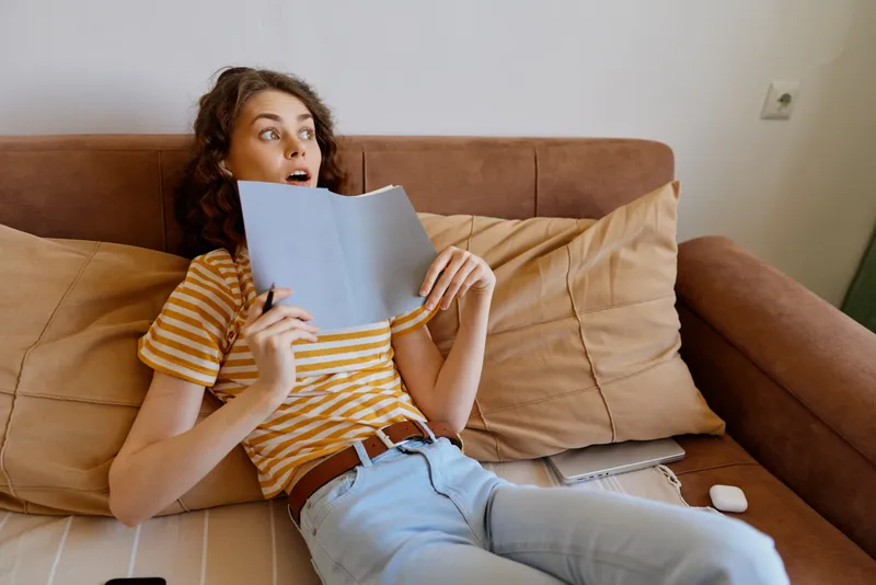Woman relaxing on a sofa at home reads a book with surprised expression, striped t shirt and jeans, cozy living room, natural light, casual lifestyle moment focused on reading