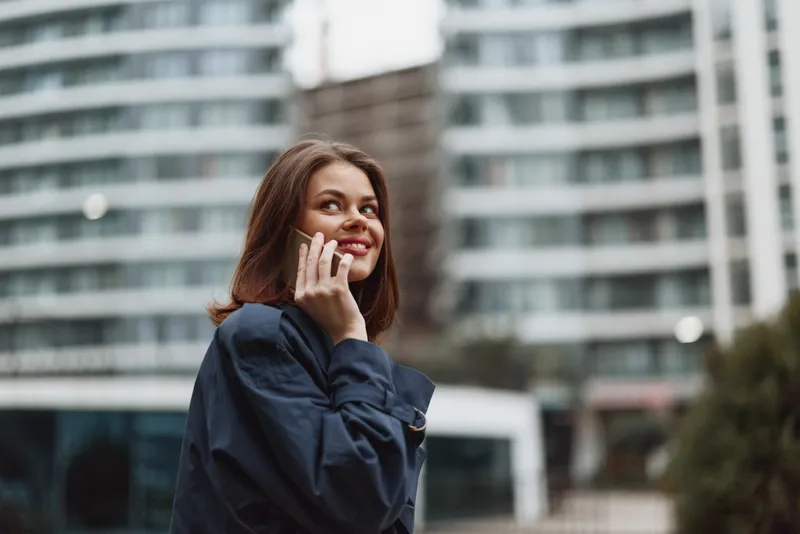 Smiling woman talking on smartphone outdoors, casual clothing, urban background, natural light, confident expression, modern lifestyle, communication moment