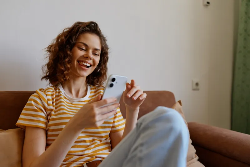 Cheerful woman indoors, relaxed on a sofa, smiling while using a smartphone, casual striped tee, soft natural light, cozy home vibe, candid moment, everyday connection, warm colors, comfortable
