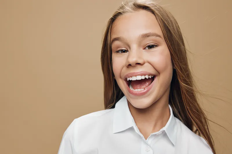 Young girl smiling and laughing in white shirt with long hair against beige background. Closeup portrait of happy female child with teeth showing and bright positive expression.