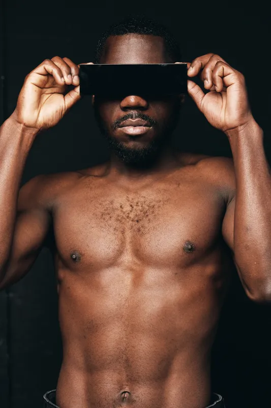 Creative conceptual portrait of shirtless African man holding smartphone horizontally over eyes with dark background, visual style emphasizing skin texture and modern technology.