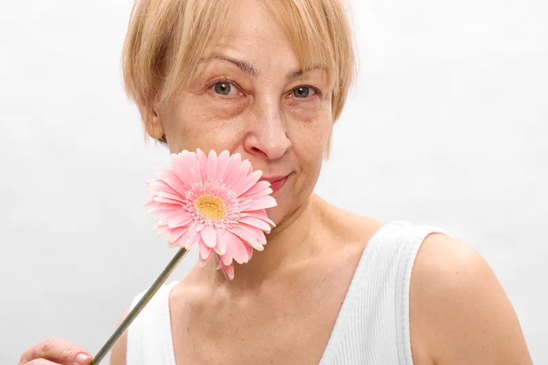 Woman holding pink gerbera flower near face, smiling softly, close up studio portrait, white background, calm mood, natural skin tones, minimal focus soft light