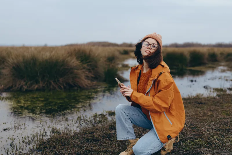 Woman lifestyle portrait outdoors by river in autumn, kneeling in grass wearing orange jacket, knit hat and scarf, relaxed smile, peaceful travel mood in daylight