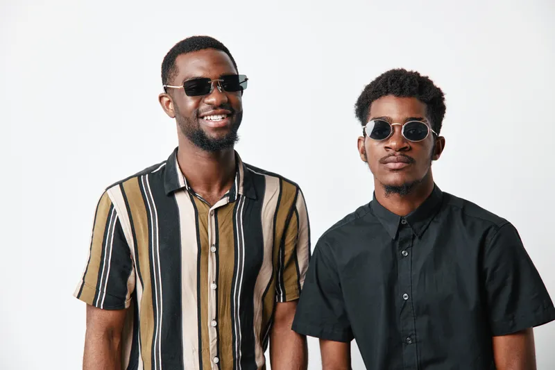 Two young black men with dark skin wearing sunglasses and casual shirts standing together on white background smiling and posing for portrait photography.