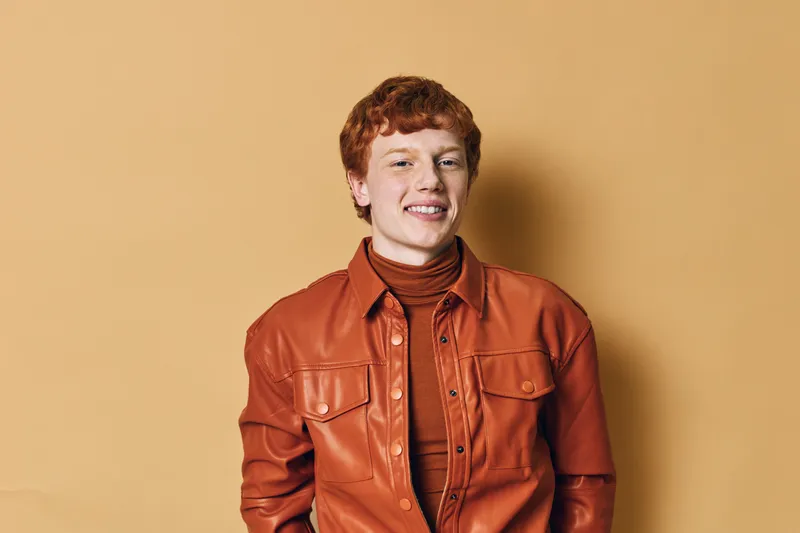 Young man with red hair wearing orange leather jacket and turtleneck sweater stands smiling against beige background in studio for portrait or fashion concept.