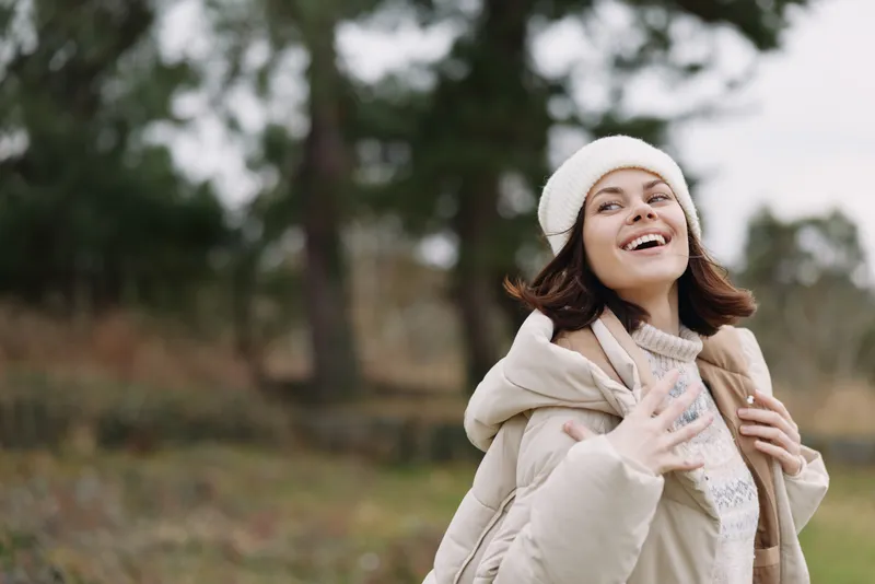 Woman outdoors in a coat and beanie, smiling in a park, candid portrait showcasing fashion and nature in cool weather, with relaxed expression and natural light.