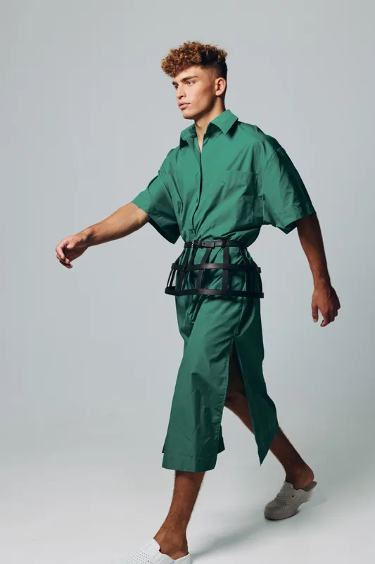 Young man walking in green oversized shirt and matching wide pants with black belt bag and white slip on shoes on gray background in studio.