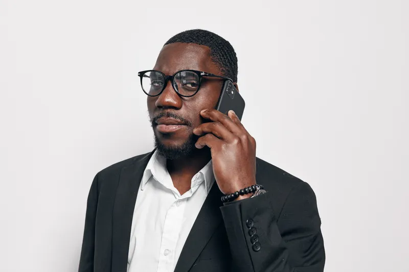 Black man with dark skin wearing glasses and black suit talking on smartphone against white background. Confident African American businessman in formal wear using mobile phone for communication.