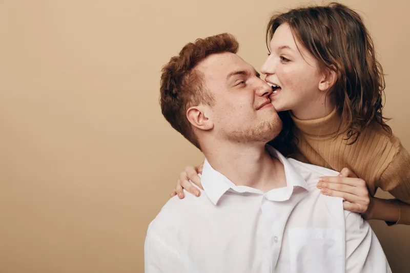 Young couple in casual clothes share intimate moment, woman gently kisses man on cheek. Warm beige background creates cozy atmosphere. Concept of love, affection, and close relationship.