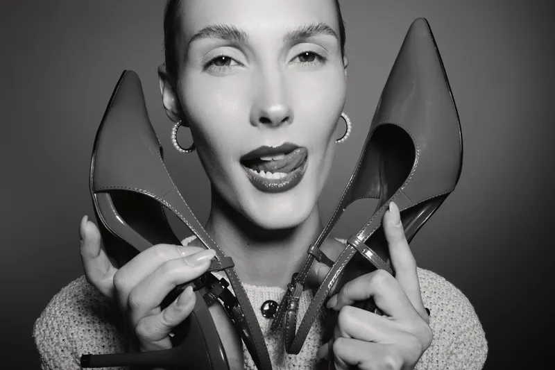 Young white woman with light skin and short hair holds high heel shoes near face, playful expression with tongue out in black and white studio portrait.