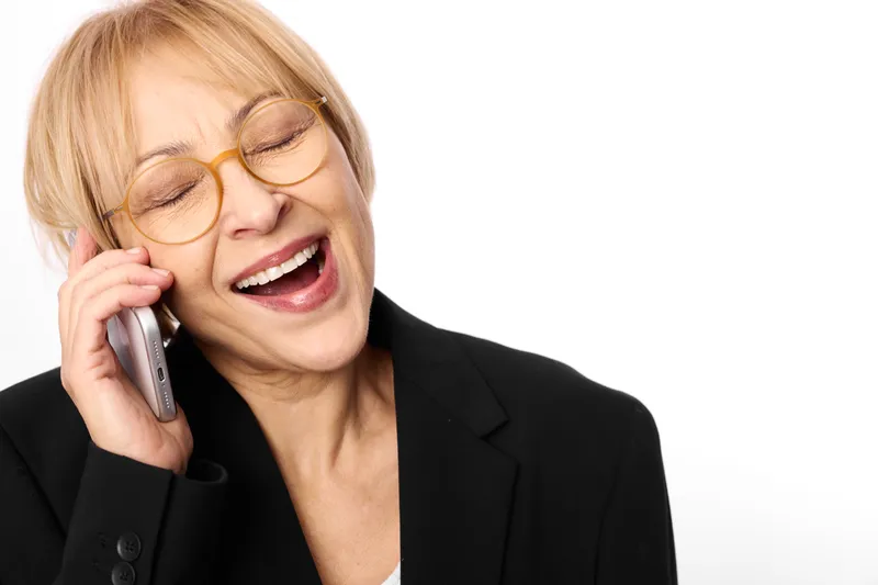 Smiling woman with glasses holding a smartphone to her ear, business jacket, close up portrait in a bright studio setting, friendly call and modern communication, calm confident mood