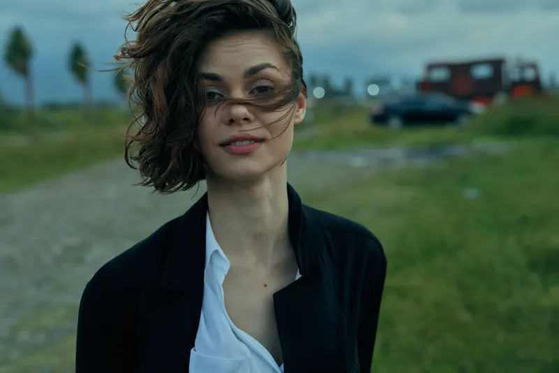 Windblown woman in blazer and white shirt outdoors, moody sky and atmospheric setting, casual fashion with confident gaze and expressive mood